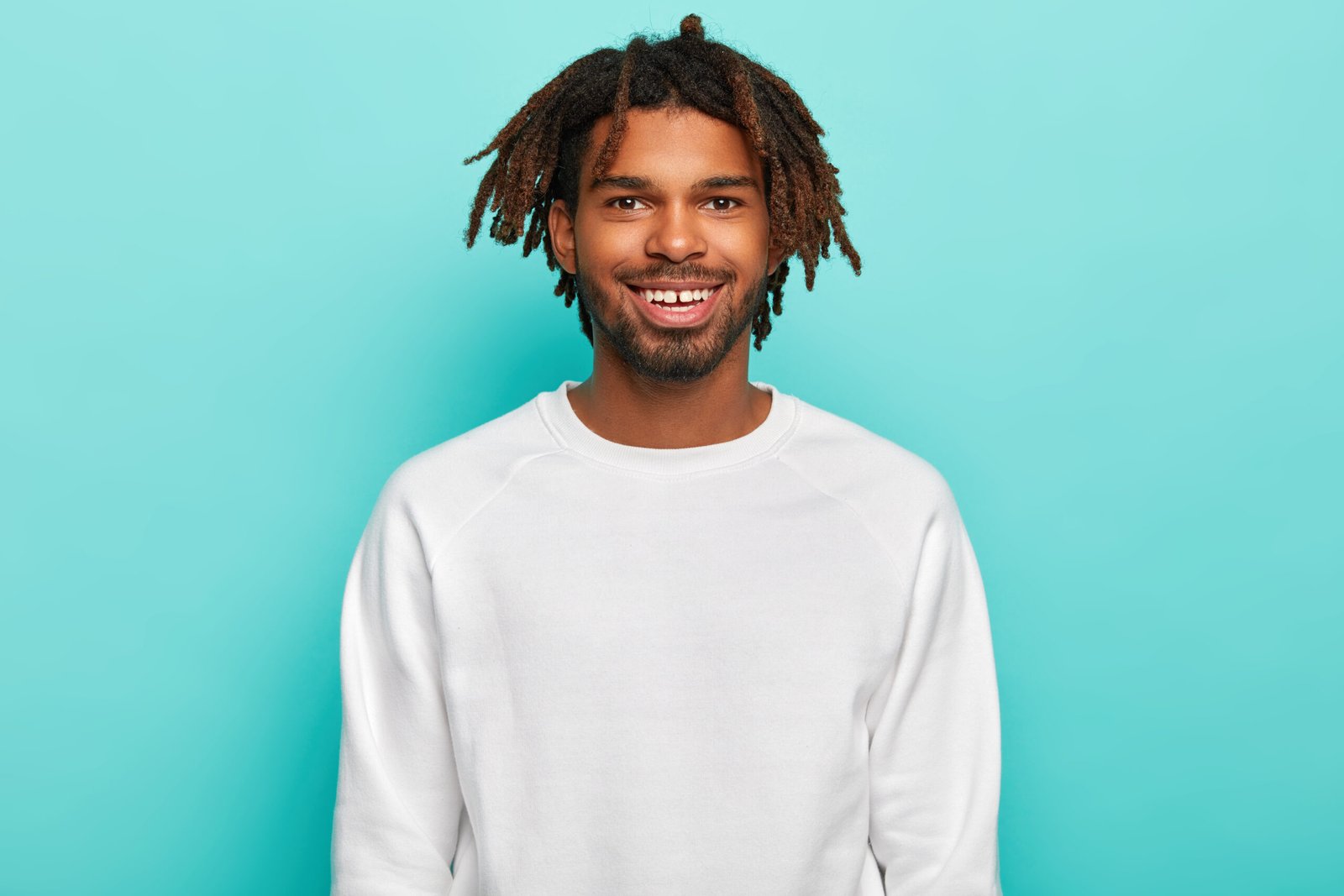 dark skinned hipster with dreadlocks, smiles broadly, shows teeth, wears white casual jumper, satisfied ater spending weekend with friends, isolated over blue background. people, ethnicity concept